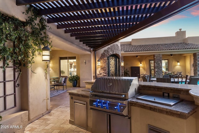 patio terrace at dusk with a pergola, ceiling fan, exterior kitchen, and a grill