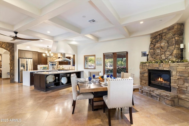 dining area with beamed ceiling, ceiling fan with notable chandelier, a stone fireplace, and coffered ceiling