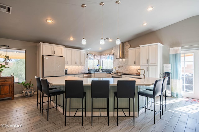 kitchen featuring visible vents, a sink, stainless steel appliances, vaulted ceiling, and wall chimney range hood