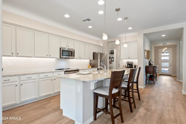 kitchen featuring appliances with stainless steel finishes, hanging light fixtures, white cabinetry, an island with sink, and light hardwood / wood-style flooring