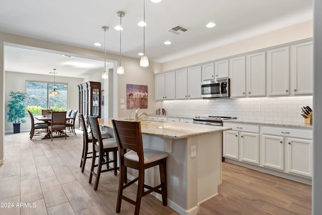 kitchen featuring hanging light fixtures, white cabinets, stainless steel appliances, light wood-type flooring, and a kitchen island with sink