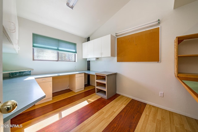 unfurnished office with lofted ceiling, baseboards, built in desk, and light wood finished floors