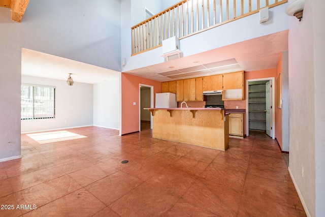 kitchen featuring baseboards, under cabinet range hood, open floor plan, freestanding refrigerator, and a towering ceiling