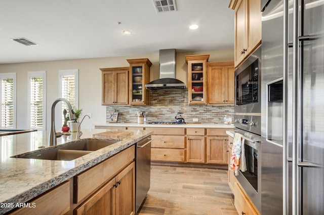 kitchen featuring wall chimney exhaust hood, light stone counters, sink, and appliances with stainless steel finishes