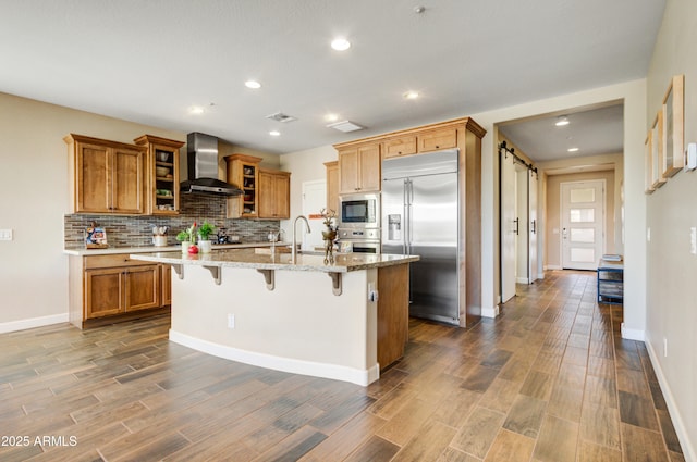 kitchen featuring built in appliances, wall chimney exhaust hood, a kitchen island with sink, and a breakfast bar area