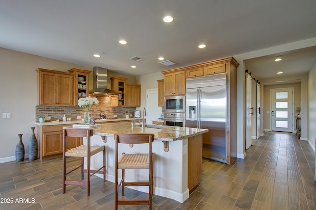 kitchen with decorative backsplash, wall chimney exhaust hood, sink, built in appliances, and a center island with sink