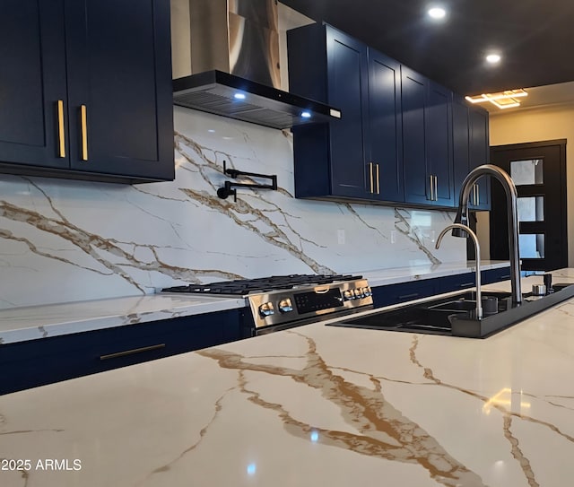 kitchen featuring wall chimney exhaust hood, blue cabinetry, decorative backsplash, light stone counters, and a sink