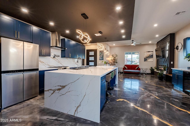kitchen with stainless steel appliances, wall chimney exhaust hood, blue cabinets, a sink, and light stone counters