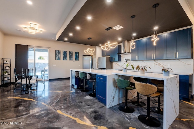 kitchen with blue cabinetry, freestanding refrigerator, wall chimney range hood, a kitchen breakfast bar, and decorative backsplash