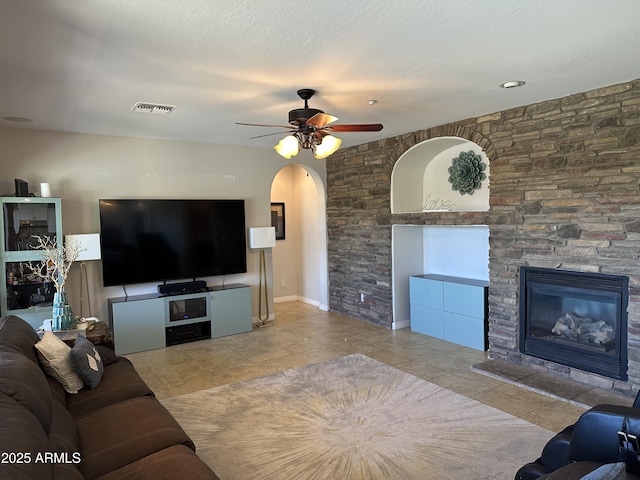 living room with a ceiling fan, visible vents, arched walkways, a stone fireplace, and tile patterned floors