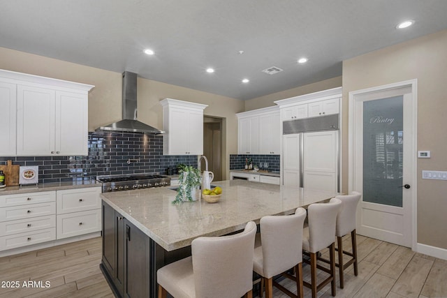 kitchen featuring wall chimney range hood, paneled built in refrigerator, white cabinets, and recessed lighting