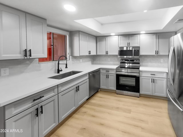 kitchen with gray cabinetry, stainless steel appliances, a sink, light wood-style floors, and a raised ceiling