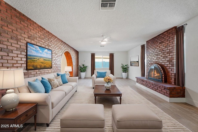 living room with ceiling fan, light hardwood / wood-style floors, a textured ceiling, brick wall, and a brick fireplace