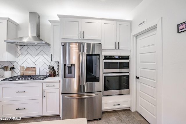 kitchen featuring wall chimney range hood, white cabinets, light countertops, stainless steel appliances, and decorative backsplash