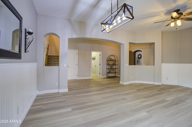 empty room featuring a ceiling fan, stairs, wainscoting, light wood-type flooring, and arched walkways