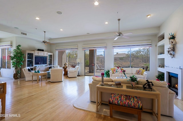 living area with light wood-style floors, a glass covered fireplace, visible vents, and plenty of natural light