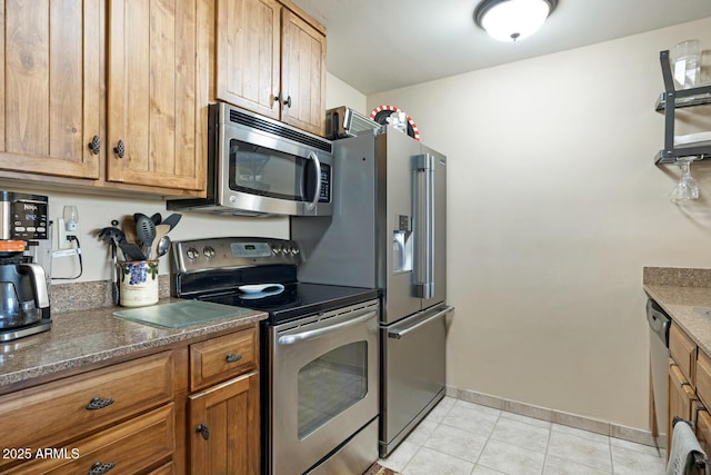 kitchen featuring baseboards, dark stone counters, light tile patterned floors, brown cabinetry, and stainless steel appliances