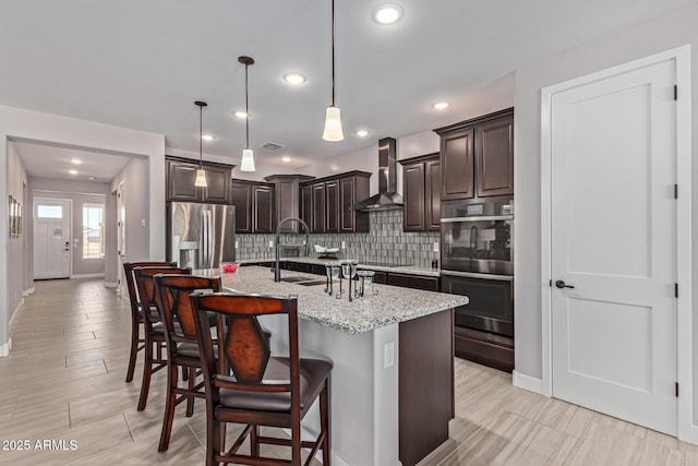 kitchen featuring decorative light fixtures, wall chimney exhaust hood, an island with sink, appliances with stainless steel finishes, and dark brown cabinets