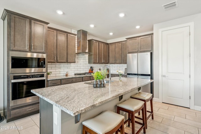 kitchen with visible vents, a sink, backsplash, appliances with stainless steel finishes, and wall chimney range hood