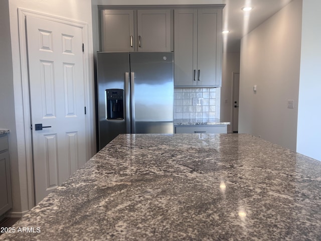 kitchen with gray cabinets, backsplash, stainless steel refrigerator with ice dispenser, and dark stone countertops