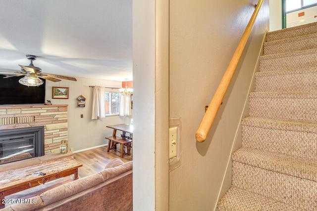 stairs featuring ceiling fan with notable chandelier, a stone fireplace, wood finished floors, and baseboards