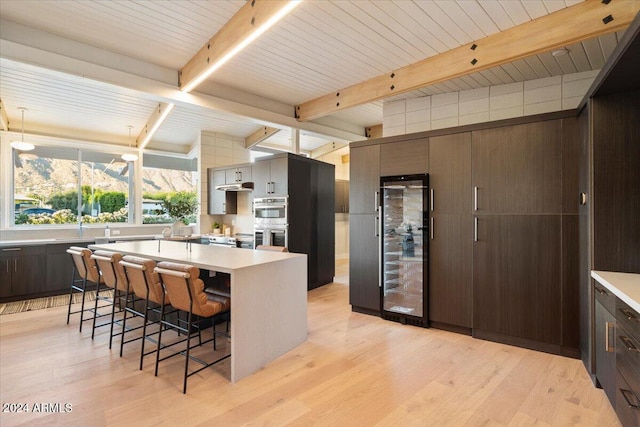 kitchen featuring double oven, a kitchen breakfast bar, beamed ceiling, beverage cooler, and light hardwood / wood-style flooring