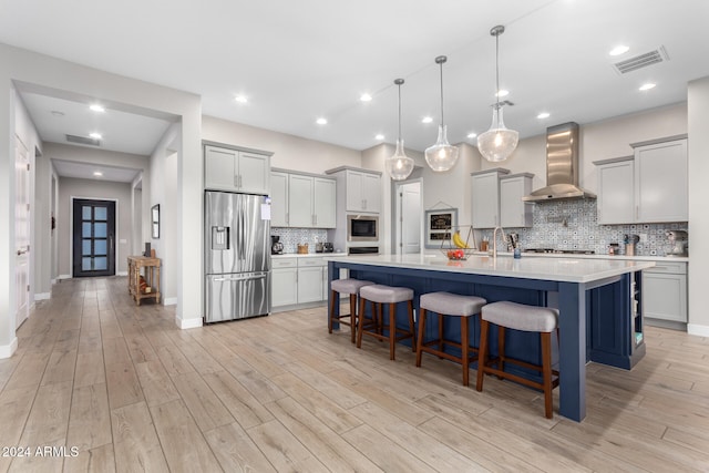 kitchen with appliances with stainless steel finishes, wall chimney exhaust hood, decorative light fixtures, a kitchen island with sink, and light wood-type flooring