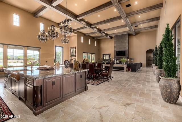 kitchen featuring arched walkways, dark brown cabinetry, light stone countertops, a large island with sink, and pendant lighting