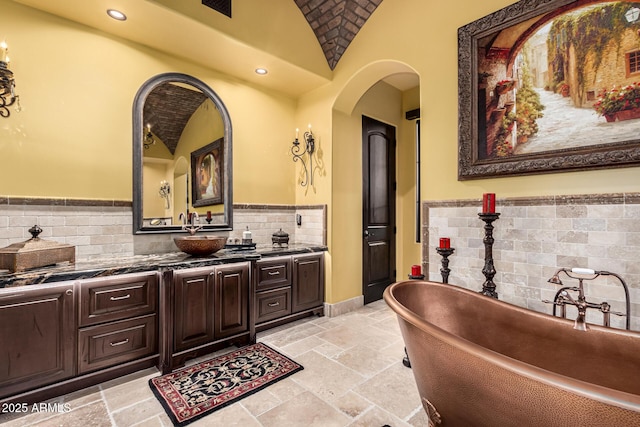 bathroom featuring lofted ceiling, recessed lighting, vanity, a soaking tub, and stone tile flooring