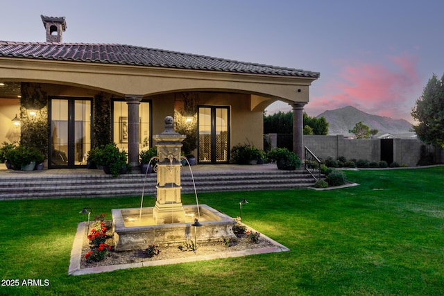 back of property at dusk with a mountain view, french doors, a lawn, and stucco siding