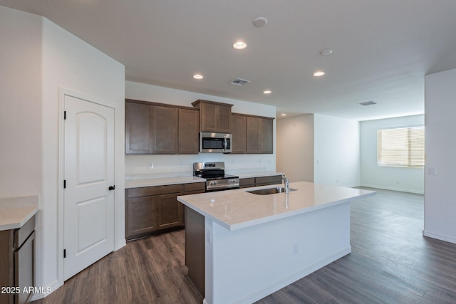 kitchen with light stone countertops, sink, dark wood-type flooring, stainless steel appliances, and a center island with sink