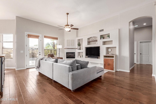 living room with built in shelves, ceiling fan, dark wood-type flooring, and a towering ceiling