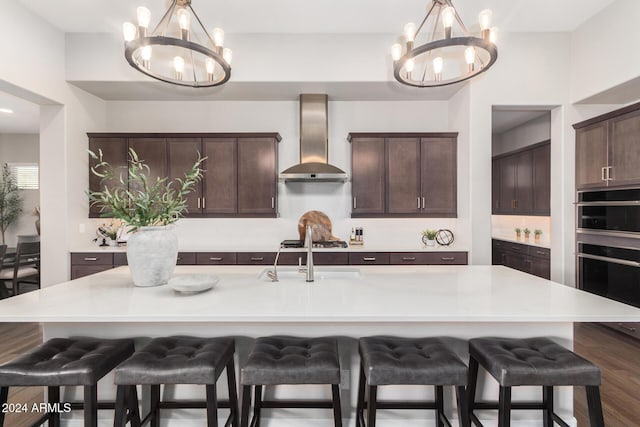 kitchen featuring wall chimney exhaust hood, dark brown cabinets, sink, a center island with sink, and an inviting chandelier