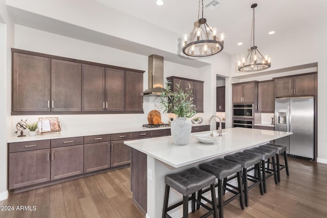 kitchen featuring a breakfast bar, a kitchen island with sink, wall chimney range hood, appliances with stainless steel finishes, and a notable chandelier