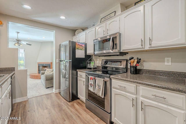 kitchen featuring light wood finished floors, a brick fireplace, stainless steel appliances, white cabinetry, and a ceiling fan