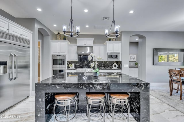 kitchen with built in appliances, a large island with sink, dark stone counters, decorative light fixtures, and decorative backsplash