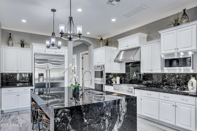 kitchen with a center island with sink, built in appliances, range hood, tasteful backsplash, and white cabinetry