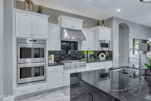 kitchen featuring ventilation hood, dark stone counters, decorative backsplash, white cabinets, and appliances with stainless steel finishes