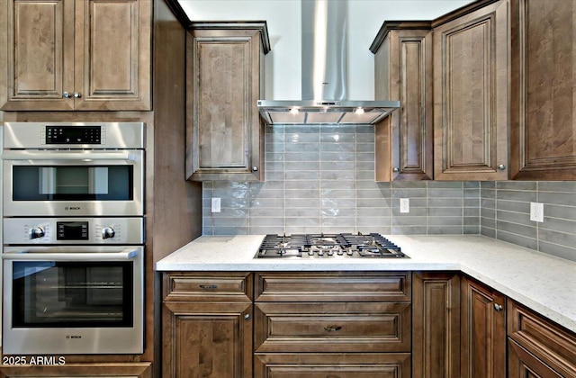 kitchen featuring decorative backsplash, dark brown cabinets, light stone counters, stainless steel appliances, and wall chimney exhaust hood
