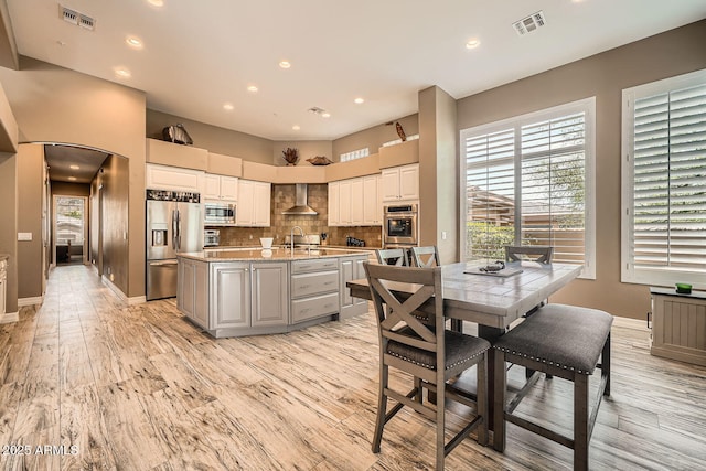 12448 N 129th Pl, Scottsdale, AZ 85259 - kitchen featuring appliances with stainless steel finishes, an island with sink, backsplash, wall chimney exhaust hood, and light wood-type flooring
