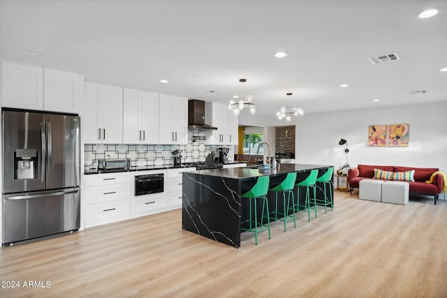 kitchen featuring stainless steel refrigerator with ice dispenser, a kitchen island with sink, wall chimney range hood, white cabinetry, and a breakfast bar area