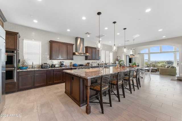 kitchen featuring light stone countertops, a spacious island, stainless steel double oven, wall chimney exhaust hood, and pendant lighting