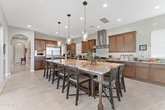 kitchen featuring light stone countertops, wall chimney range hood, hanging light fixtures, a large island, and stainless steel appliances