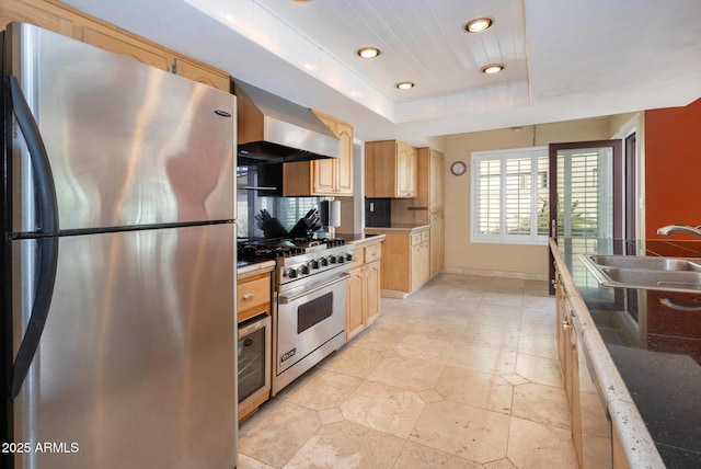 kitchen featuring wall chimney range hood, sink, a tray ceiling, light brown cabinets, and stainless steel appliances