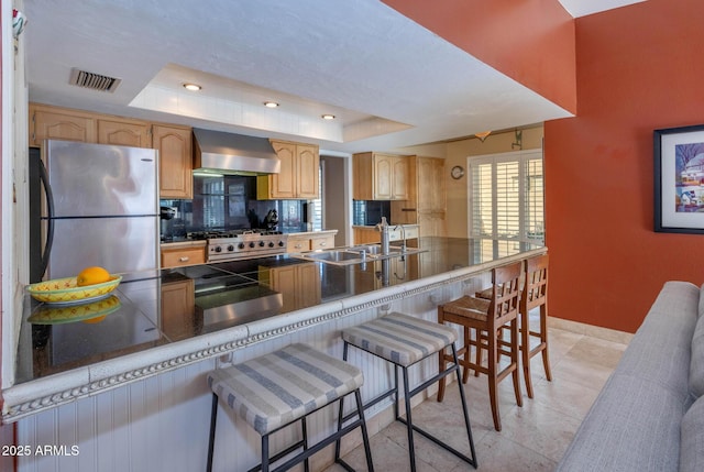 kitchen with kitchen peninsula, a breakfast bar area, a tray ceiling, wall chimney range hood, and sink