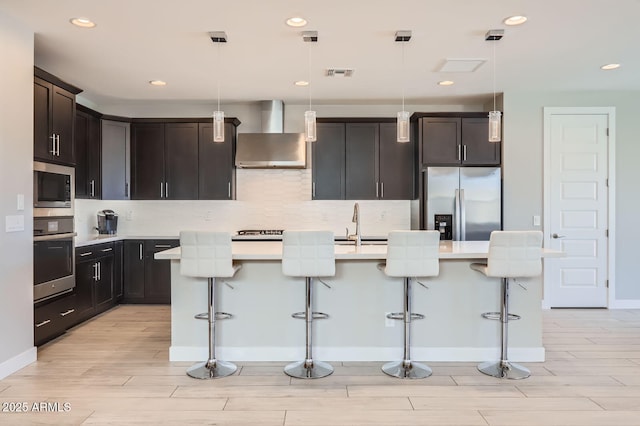 kitchen featuring visible vents, wall chimney range hood, an island with sink, light countertops, and appliances with stainless steel finishes