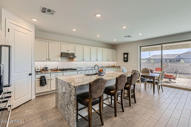 kitchen featuring stainless steel gas cooktop, a kitchen island with sink, light stone countertops, and light wood-type flooring