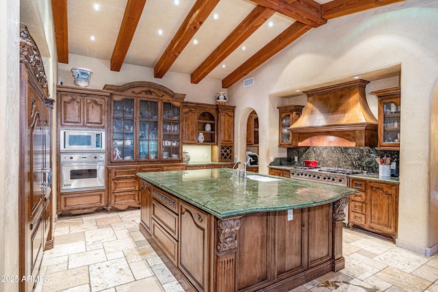 kitchen featuring custom exhaust hood, tasteful backsplash, appliances with stainless steel finishes, an island with sink, and beam ceiling