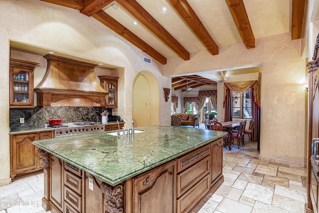 kitchen featuring beamed ceiling, sink, custom exhaust hood, range, and a center island with sink