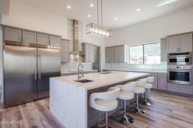 kitchen with wall chimney range hood, light stone counters, light hardwood / wood-style floors, a kitchen island with sink, and appliances with stainless steel finishes